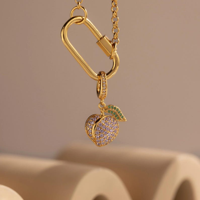 May include: A gold-colored necklace featuring a large carabiner-style clasp and a peach pendant. The peach pendant is encrusted with purple and clear stones, with a green leaf accent. The necklace is suspended against a neutral background.