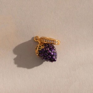 May include: A gold-colored charm shaped like a bunch of grapes. The grapes are a deep purple color with a textured surface. The charm has a gold-colored bail with clear stones and green leaves.