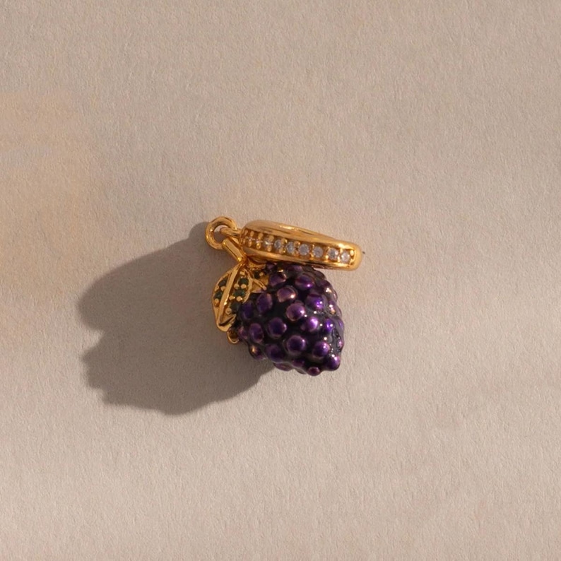 May include: A gold-colored charm shaped like a bunch of grapes. The grapes are a deep purple color with a textured surface. The charm has a gold-colored bail with clear stones and green leaves.