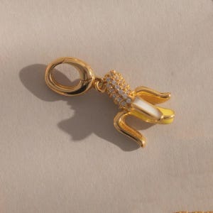May include: A gold-colored charm in the shape of a peeled banana. The banana has yellow and white enamel detailing, with a section of the banana covered in small, clear crystals. The charm has a gold clasp for attaching to a bracelet or necklace.