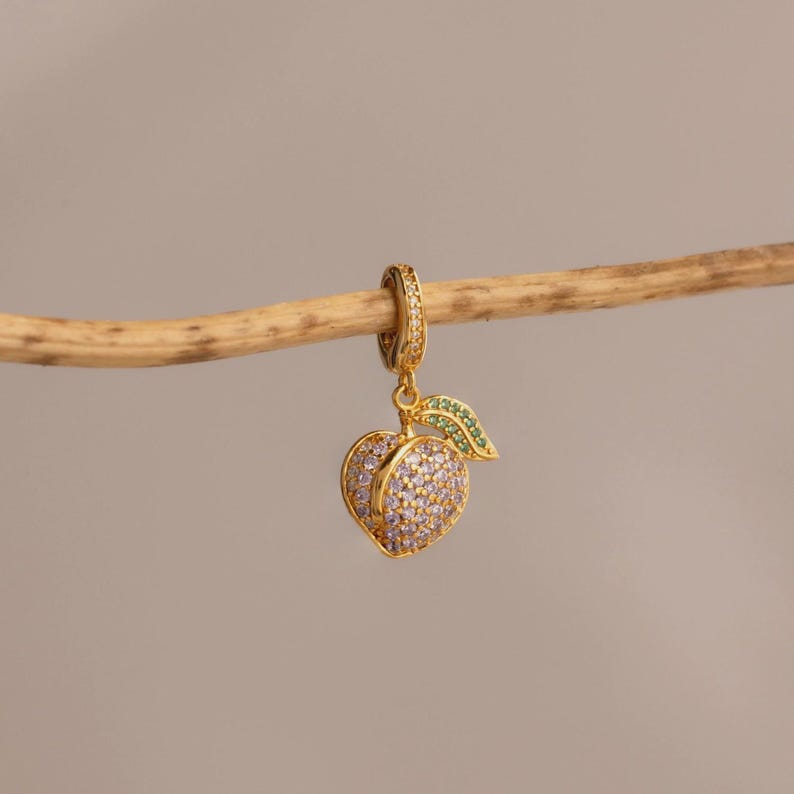 May include: A gold-colored charm in the shape of a peach, adorned with purple and clear gemstones. A green gemstone leaf is attached to the peach. The charm is suspended from a gold-colored hoop, and is hanging from a wooden stick.