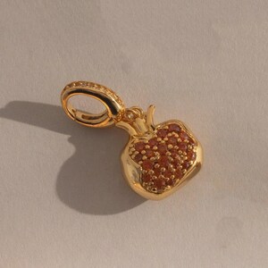 May include: A gold-colored pendant in the shape of a pomegranate. The fruit is filled with small, red-brown stones. The pendant has a loop at the top for a chain and is set against a neutral background.