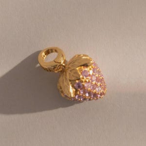 May include: A gold-colored charm in the shape of a strawberry. The strawberry is encrusted with small, pink gemstones. The charm has a loop for attaching to a bracelet or necklace. The leaves are detailed.