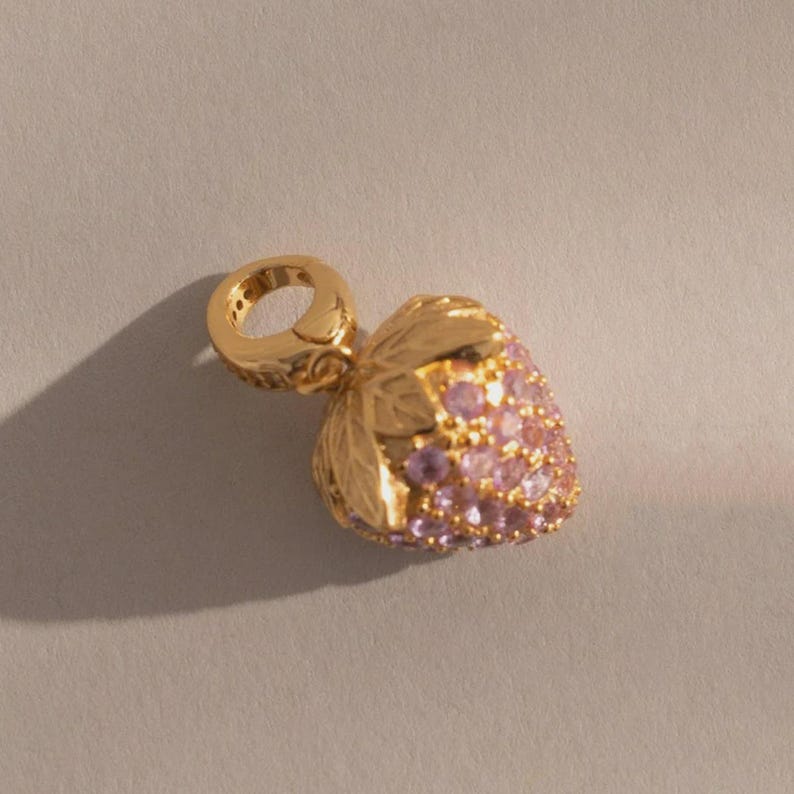 May include: A gold-colored charm in the shape of a strawberry. The strawberry is encrusted with small, pink gemstones. The charm has a loop for attaching to a bracelet or necklace. The leaves are detailed.