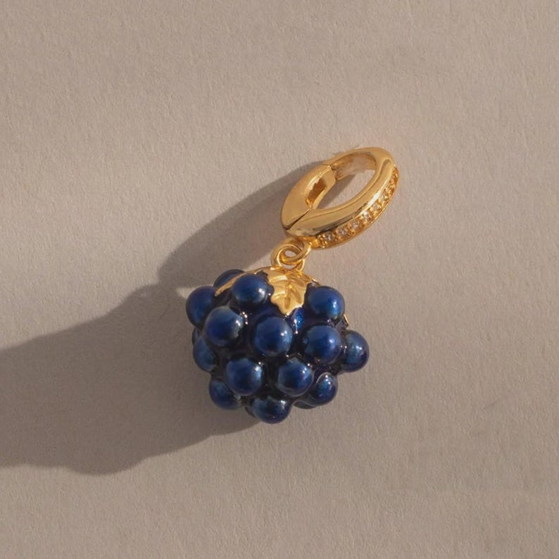 May include: A gold-colored earring with a cluster of dark blue, round beads resembling grapes. The earring has a hinged clasp and a small gold leaf detail. The beads are clustered together, creating a three-dimensional effect.