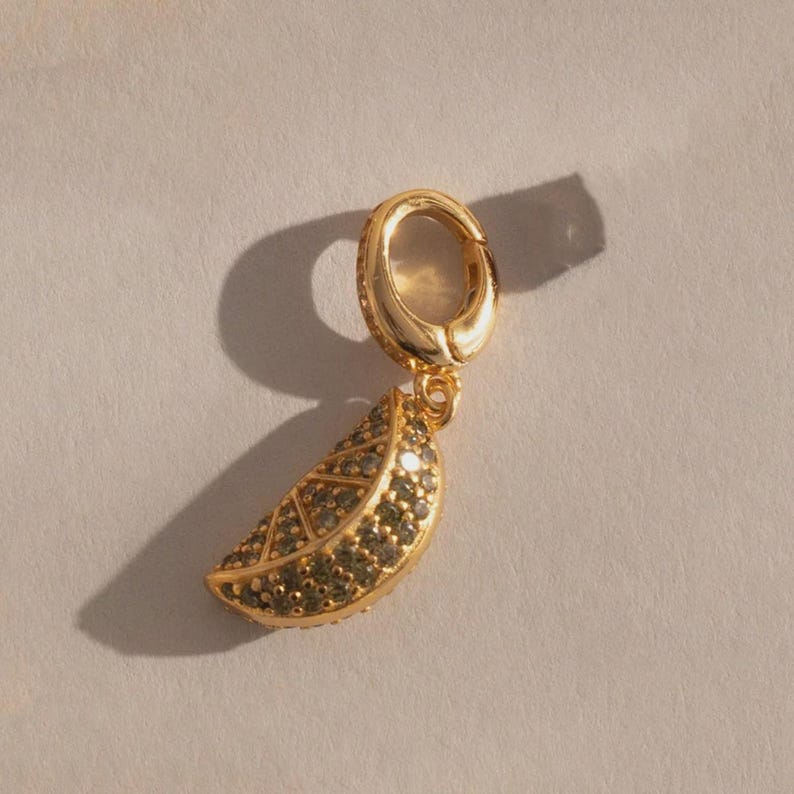 May include: A gold-colored charm with a leaf design, encrusted with small, dark green stones. The charm is attached to a gold hoop. The leaf charm has a textured surface and is casting a shadow on a light-colored background.