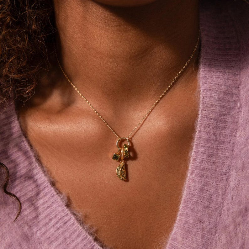 May include: A delicate gold necklace featuring a charm cluster with green and yellow gemstone accents. The pendant includes a crescent shape and other small, round charms. The necklace is worn against a soft, light purple sweater.