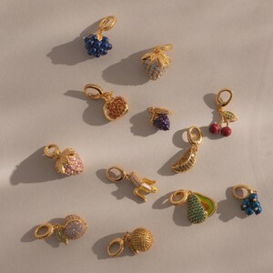 May include: A collection of gold-toned fruit charms, including grapes, strawberries, apples, cherries, bananas, and avocados. Each charm is embellished with colorful gemstones, creating a sparkling and detailed appearance. The charms are designed with a loop for easy attachment.