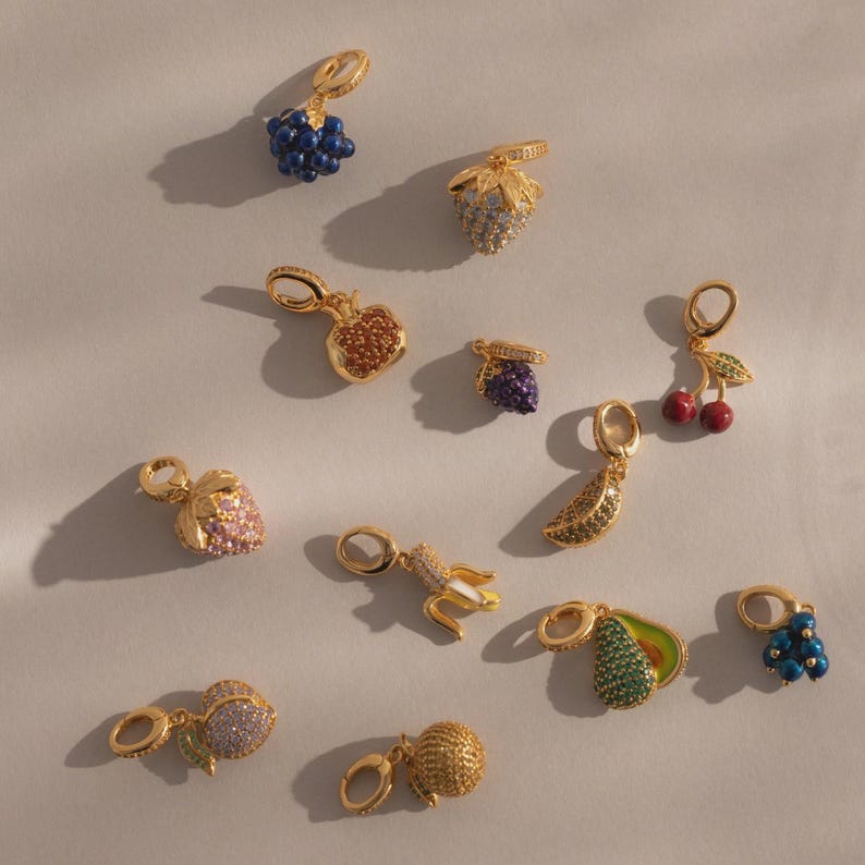 May include: A collection of gold-toned fruit charms, including grapes, strawberries, apples, cherries, bananas, and avocados. Each charm is embellished with colorful gemstones, creating a sparkling and detailed appearance. The charms are designed with a loop for easy attachment.