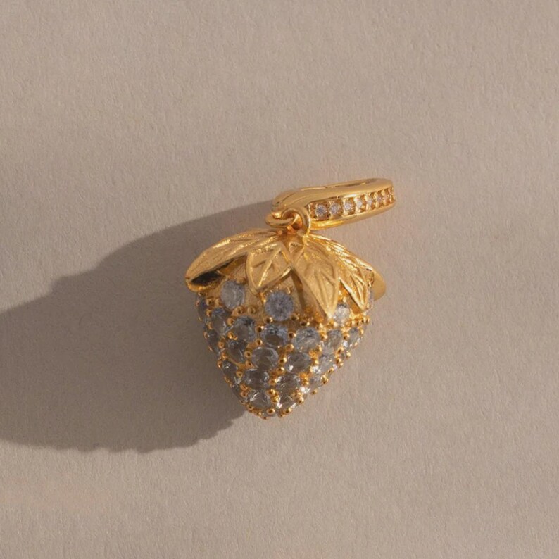 May include: A gold-colored strawberry pendant with a leaf-shaped top and a loop for a chain. The strawberry is encrusted with small, light blue gemstones. The loop is adorned with small, clear gemstones.