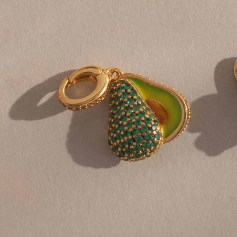 May include: Gold-toned avocado charm earrings. One half of the avocado is encrusted with green gemstones, the other half is green enamel with a brown pit. The earrings have a gold clasp.
