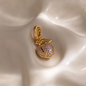 May include: A gold-colored peach charm pendant with a loop for hanging. The peach is covered in small, light purple and clear stones. A green leaf with small green stones is attached to the peach. The charm is on a white surface.