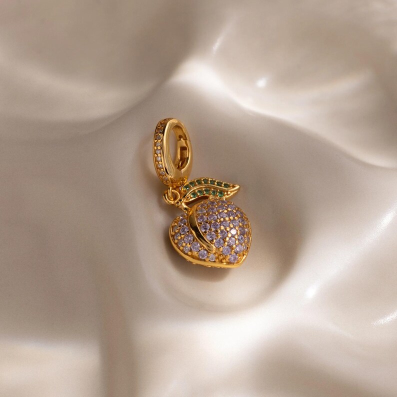May include: A gold-colored peach charm pendant with a loop for hanging. The peach is covered in small, light purple and clear stones. A green leaf with small green stones is attached to the peach. The charm is on a white surface.
