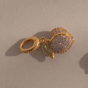 May include: A gold-colored charm featuring a peach design. The peach is covered in small, light purple stones, with green stones on the leaves. The charm includes a gold clasp for attachment.