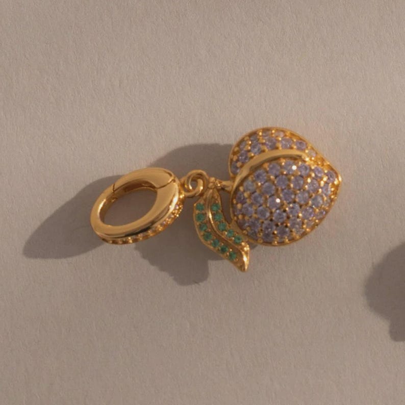 May include: A gold-colored charm featuring a peach design. The peach is covered in small, light purple stones, with green stones on the leaves. The charm includes a gold clasp for attachment.