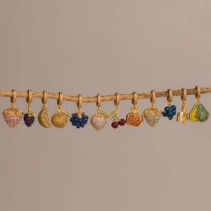 May include: A collection of gold-colored hoop earrings, each adorned with a different fruit charm. The charms include a strawberry, grapes, a lemon slice, a golden apple, blueberries, a heart-shaped apple, cherries, a pomegranate, a banana, and an avocado.
