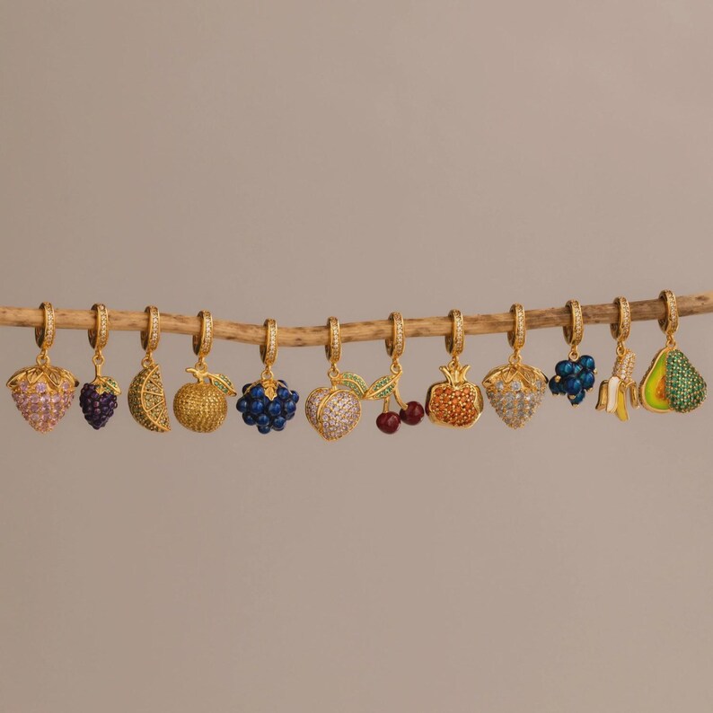 May include: A collection of gold-colored hoop earrings, each adorned with a different fruit charm. The charms include a strawberry, grapes, a lemon slice, a golden apple, blueberries, a heart-shaped apple, cherries, a pomegranate, a banana, and an avocado.