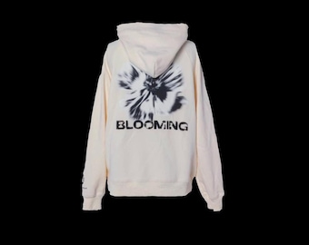 May include: Cream-colored hoodie with a black and white floral design on the back. The word "BLOOMING" is printed in black below the graphic. The hoodie has a hood and long sleeves.