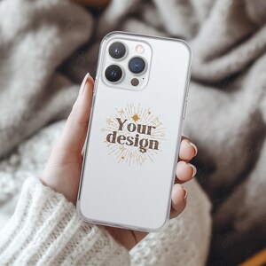 Phone case mockup of a clear phone case held by a girl's hand that's wearing a wool white sweater, in front of a cozy blanket. Works in Photoshop, Photopea and Canva.