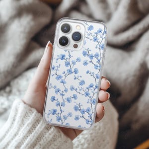 Phone case mockup of a clear phone case held by a girl's hand that's wearing a wool white sweater, in front of a cozy blanket. Works in Photoshop, Photopea and Canva.