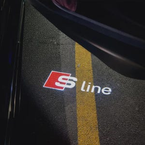 Puede incluir: Logotipo S line iluminado proyectado sobre asfalto. El logotipo presenta un diseño rojo y blanco, con el texto "S line" en blanco. Una franja amarilla recorre verticalmente la imagen, con un marco oscuro de puerta de vehículo en el fondo.