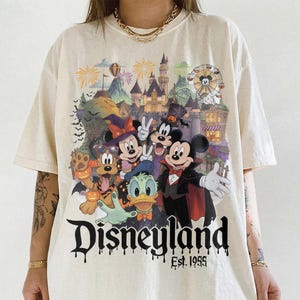 May include: An off-white short-sleeve t-shirt featuring a colorful graphic of Disney characters in Halloween costumes in front of a castle. The text Disneyland Est. 1955 is printed in a black dripping font below the characters. The shirt has a crew neck.