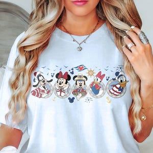 May include: A light blue t-shirt featuring a nautical-themed design with cartoon characters. The design includes images of Goofy, Minnie Mouse, Mickey Mouse, Daisy Duck, and Donald Duck in circular frames. The shirt has short sleeves.