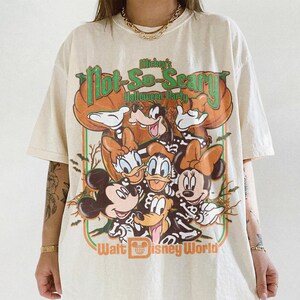May include: An off-white short-sleeve t-shirt featuring a graphic print of Mickey Mouse, Minnie Mouse, Donald Duck, Daisy Duck, and Goofy wearing skeleton costumes. The text reads Mickey's Not-So-Scary Halloween Party and Walt Disney World.