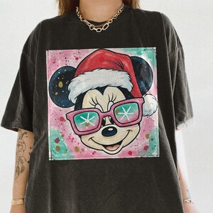 May include: Dark gray t-shirt featuring a colorful graphic of Mickey Mouse wearing a Santa hat and pink sunglasses with snowflake lenses. The graphic is set against a pink and teal speckled background. The shirt has short sleeves and a relaxed fit.