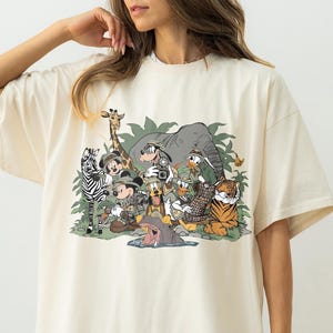 K&ouml;nnte beinhalten: Cremefarbenes, &uuml;bergro&szlig;es T-Shirt mit einem farbenfrohen Grafikdesign von Zeichentierfiguren, darunter ein Zebra, eine Giraffe, ein Elefant, ein Tiger und Mickey Mouse. Das Design ist vor einem Hintergrund aus gr&uuml;nem Laub angeordnet.