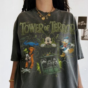 May include: A charcoal gray oversized t-shirt featuring a graphic print of the Tower of Terror with Mickey Mouse, Goofy, and Donald Duck. The design includes green lightning accents and the text Tower of Terror in a stylized font across the chest.