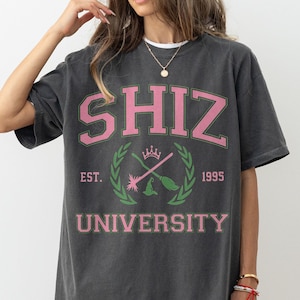 Pu&ograve; includere: T-shirt grigio scuro con la scritta "SHIZ UNIVERSITY" in rosa e verde. La maglietta presenta un design in stile collegiale con uno stemma che include scope incrociate, una corona e l'anno 1995.
