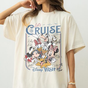 May include: Cream-colored oversized t-shirt featuring a graphic of Disney characters, including Mickey and Minnie Mouse, Donald Duck, and Goofy. The shirt has the text "Let's Cruise" and "Disney Wish" in blue.