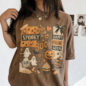 K&ouml;nnte beinhalten: Braunes Halloween-T-Shirt mit einem grafischen Design mit K&uuml;rbissen, Geistern, einem Hexenhut und den Worten "Spooky Boo! Happy Halloween". Das Design umfasst Sterne, Blumen und einen Cupcake.