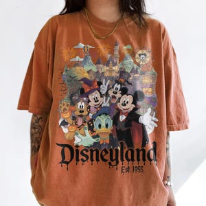 May include: An oversized, washed-out orange t-shirt featuring a graphic of Mickey Mouse, Minnie Mouse, Donald Duck, and Pluto in Halloween costumes in front of a castle. The text reads Disneyland Est. 1955 in a black, dripping, gothic-style font.