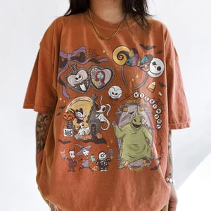 May include: Burnt orange t-shirt with a colourful graphic from The Nightmare Before Christmas. The design includes Jack Skellington, Sally, Oogie Boogie, and other characters. The shirt has a relaxed fit and short sleeves.