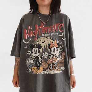 May include: Dark gray t-shirt featuring a Halloween-themed graphic. The design includes Mickey and Minnie Mouse dressed as skeletons, the words "Nightmare on Main Street," bats, and a full moon. The shirt has a vintage, oversized fit.
