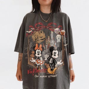 May include: Dark gray t-shirt featuring a Halloween-themed graphic. The design includes Mickey and Minnie Mouse dressed as skeletons, a haunted castle, and the words "Nightmare on Main Street."
