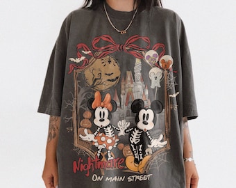 Puede incluir: Camiseta gris oscuro con un diseño temático de Halloween. El diseño incluye a Mickey y Minnie Mouse vestidos de esqueletos, un castillo embrujado y la frase "Nightmare on Main Street".