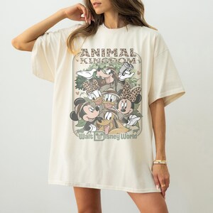 May include: Cream-colored oversized t-shirt featuring a graphic of Disney characters, including Mickey and Minnie Mouse, Donald Duck, and Goofy. The shirt has the text "ANIMAL KINGDOM" and "Walt Disney World" in a vintage style.
