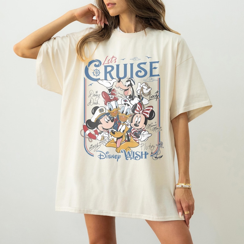 May include: Cream-colored oversized t-shirt featuring a graphic of Disney characters, including Mickey and Minnie Mouse, Donald Duck, and Goofy. The shirt has the text "Let's Cruise" and "Disney Wish" in blue.