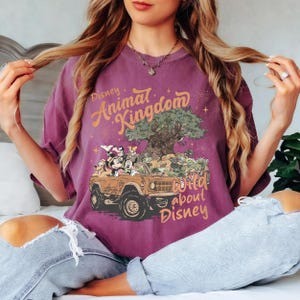 May include: A purple t-shirt featuring the text "Disney Animal Kingdom" in orange script. The graphic shows Disney characters in a jeep with a tree and the text "Wild about Disney".