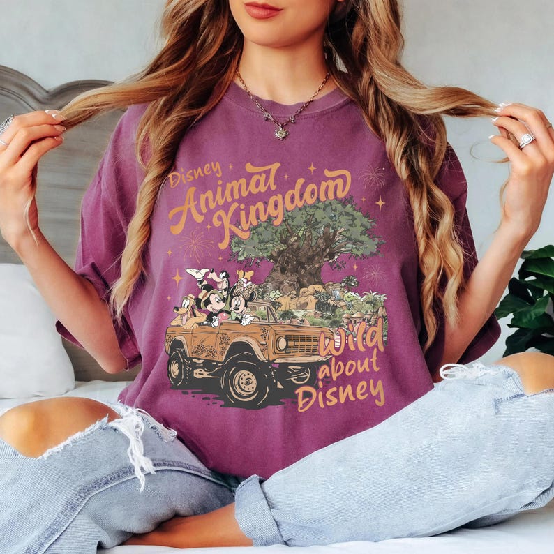 May include: A purple t-shirt featuring the text "Disney Animal Kingdom" in orange script. The graphic shows Disney characters in a jeep with a tree and the text "Wild about Disney".
