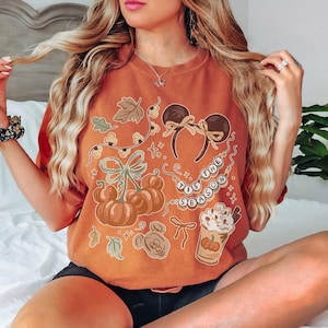 May include: A burnt orange t-shirt featuring a graphic print with mouse ears, pumpkins, a coffee cup, and the text 'TIS THE SEASON'. The design includes autumn leaves, bows, and string lights in shades of brown, cream, and muted green.