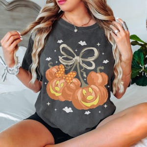 May include: A charcoal grey short-sleeve t-shirt featuring a graphic of two orange pumpkin jack-o'-lanterns shaped like mouse heads. One pumpkin wears a yellow and orange polka-dot bow. The design includes small white stars and bat silhouettes.