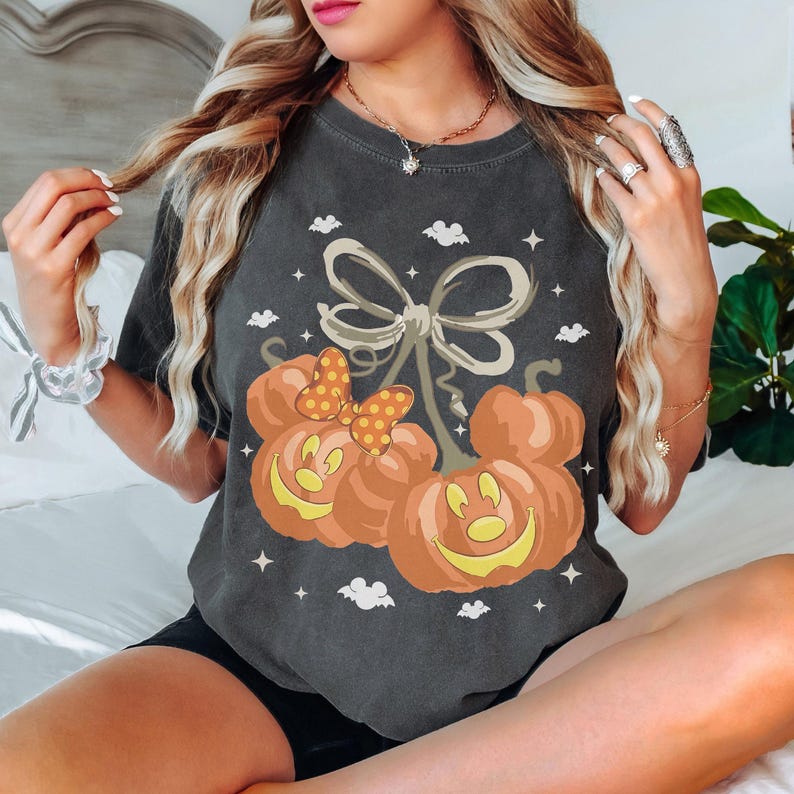 May include: A charcoal grey short-sleeve t-shirt featuring a graphic of two orange pumpkin jack-o'-lanterns shaped like mouse heads. One pumpkin wears a yellow and orange polka-dot bow. The design includes small white stars and bat silhouettes.