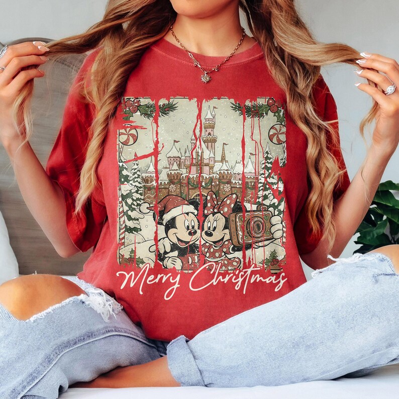 May include: Red t-shirt featuring a graphic of Mickey and Minnie Mouse in front of a castle, with the words "Merry Christmas" below. The design includes Christmas trees, candy canes, and holly. The shirt is a solid red color.