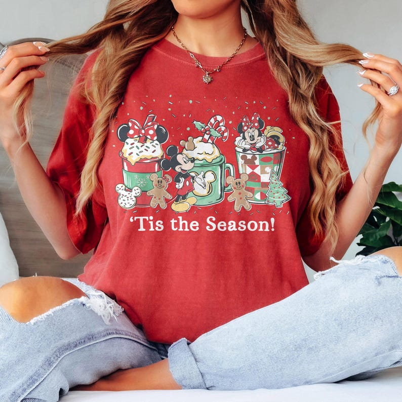 Puede incluir: Camiseta roja con un dise&ntilde;o navide&ntilde;o que presenta a Mickey y Minnie Mouse, hombres de jengibre y la frase "'Tis the Season!". El dise&ntilde;o incluye golosinas y decoraciones festivas.