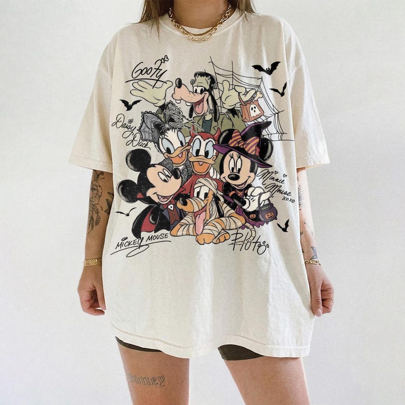 Może przedstawiać: Kremowy t-shirt z kr&oacute;tkim rękawem z nadrukiem graficznym przedstawiającym postacie Disneya w kostiumach na Halloween, w tym Myszkę Miki, Myszkę Minnie, Goofy'ego, Kaczora Donalda, Daisy i Pluto, z napisami oraz nietoperzami.