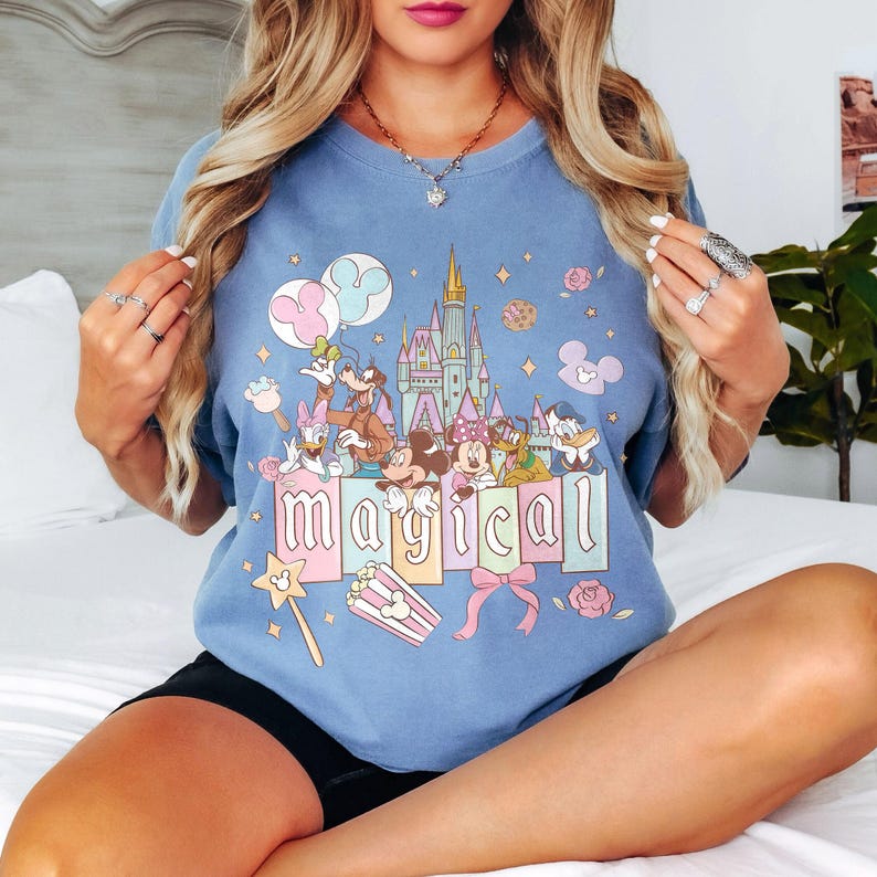 May include: A light blue t-shirt featuring a colorful graphic of Disney characters, a castle, and the word "magical". The design includes Mickey and Minnie Mouse, Donald Duck, Goofy, and other characters. The shirt has a crew neck and short sleeves.